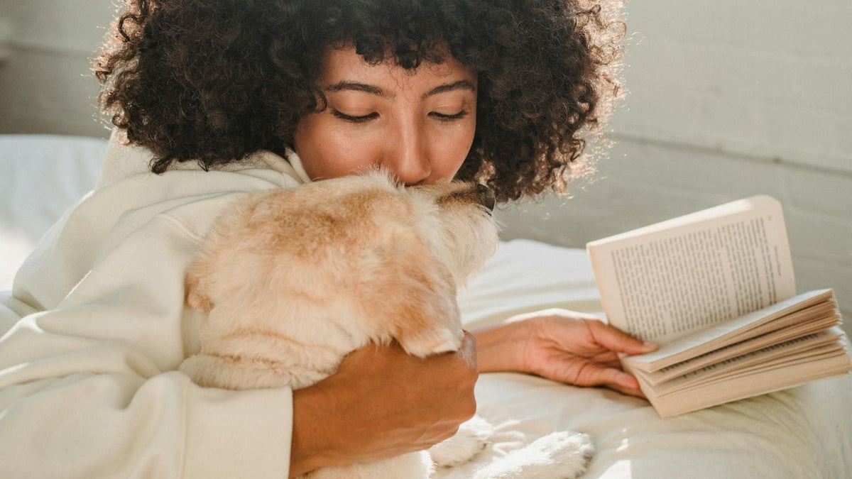 A person sitting on the bed, sharing a heartfelt hug with their dog while holding a book, representing the bond of learning together.