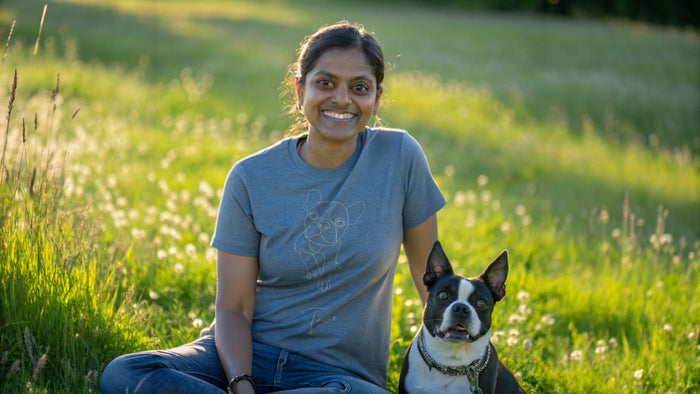A woman in a Comfort Colors t-shirt with a minimal line art drawing of a Boston Terrier sits in a sunny summer field, petting her real Boston Terrier.