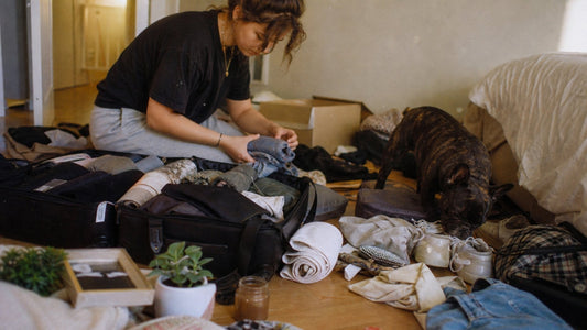 A woman packing clothes into a suitcase in a bedroom, with a dog standing nearby and various bags and clothing scattered on the floor.