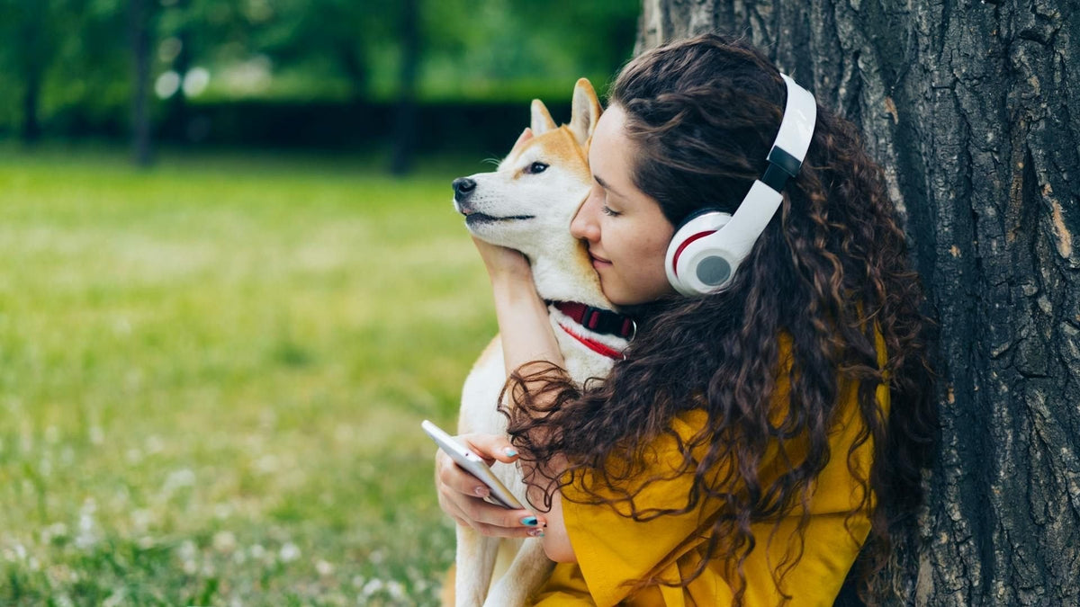 A young woman wearing headphones sits under a tree, lovingly hugging her happy golden retriever while they share a quiet moment listening to music.